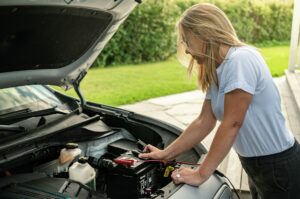 Kvinne står foran en bil med åpent panser og putter klemmer på 12-voltsbatteriet for å lade det.