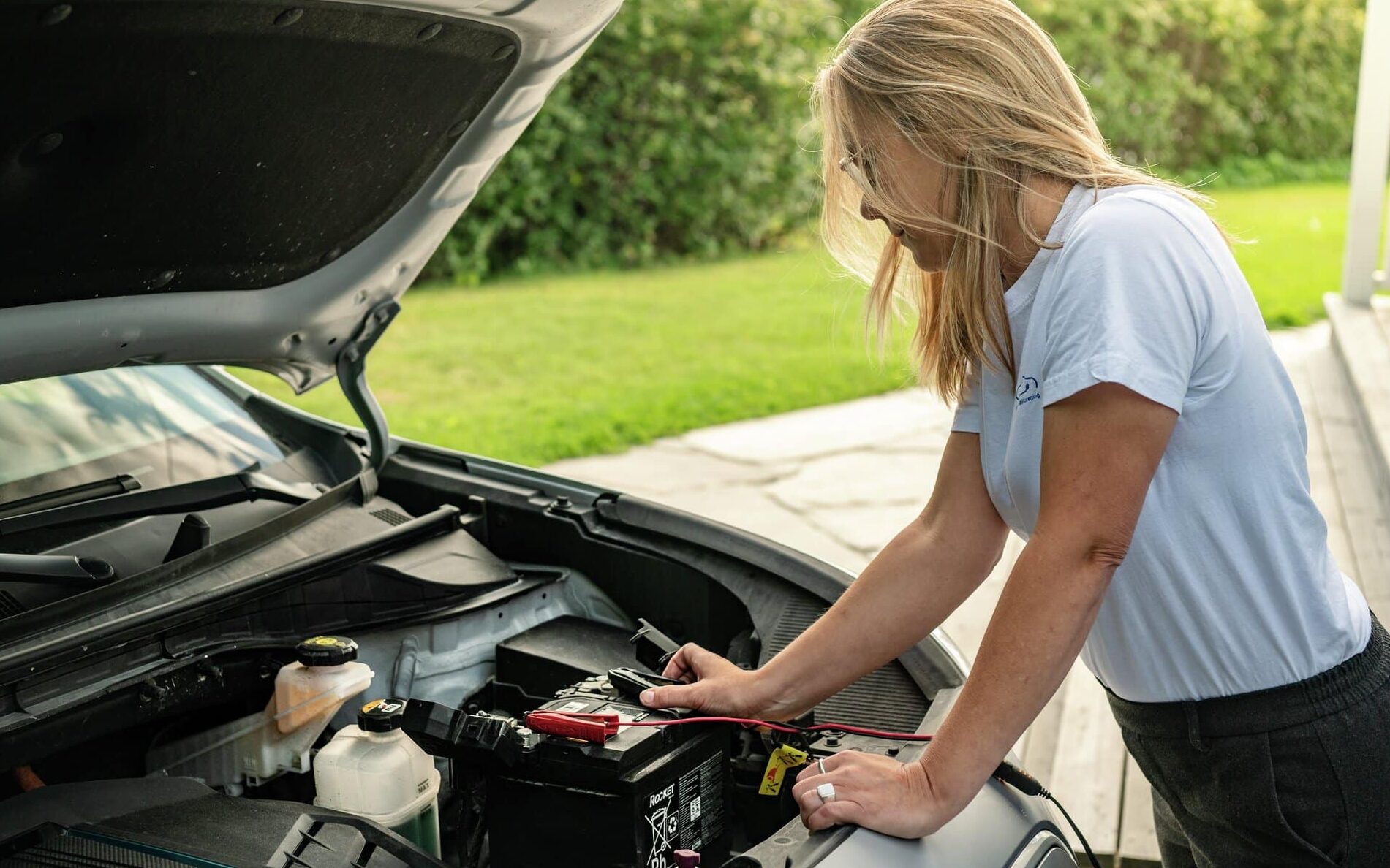 Kvinne står foran en bil med åpent panser og putter klemmer på 12-voltsbatteriet for å lade det.